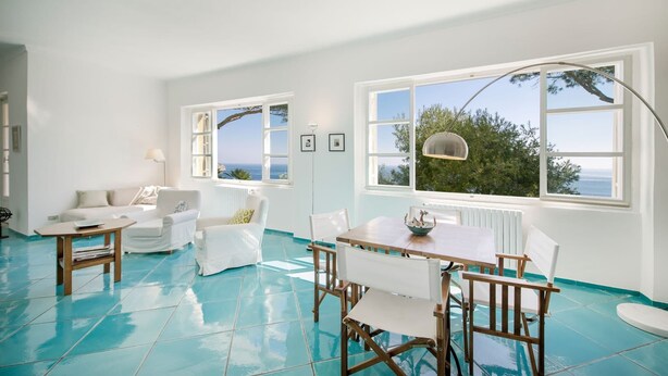 A bright room with a large window overlooking the sea, a white sofa, a wooden table, and a blue-tiled floor. - Fisherman's house in the harbor of Procida (Procida)