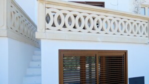 Un bâtiment à la façade blanche, doté d’une fenêtre en bois avec des stores horizontaux et d’une balustrade décorative ornée de motifs circulaires.
