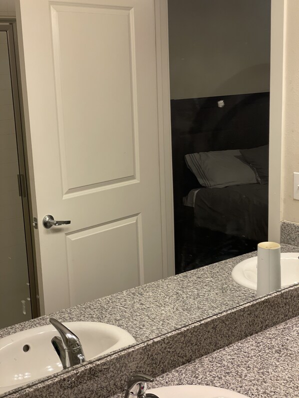 A bathroom with a white door, a sink with a faucet, and a mirror reflecting a bed with pillows.
