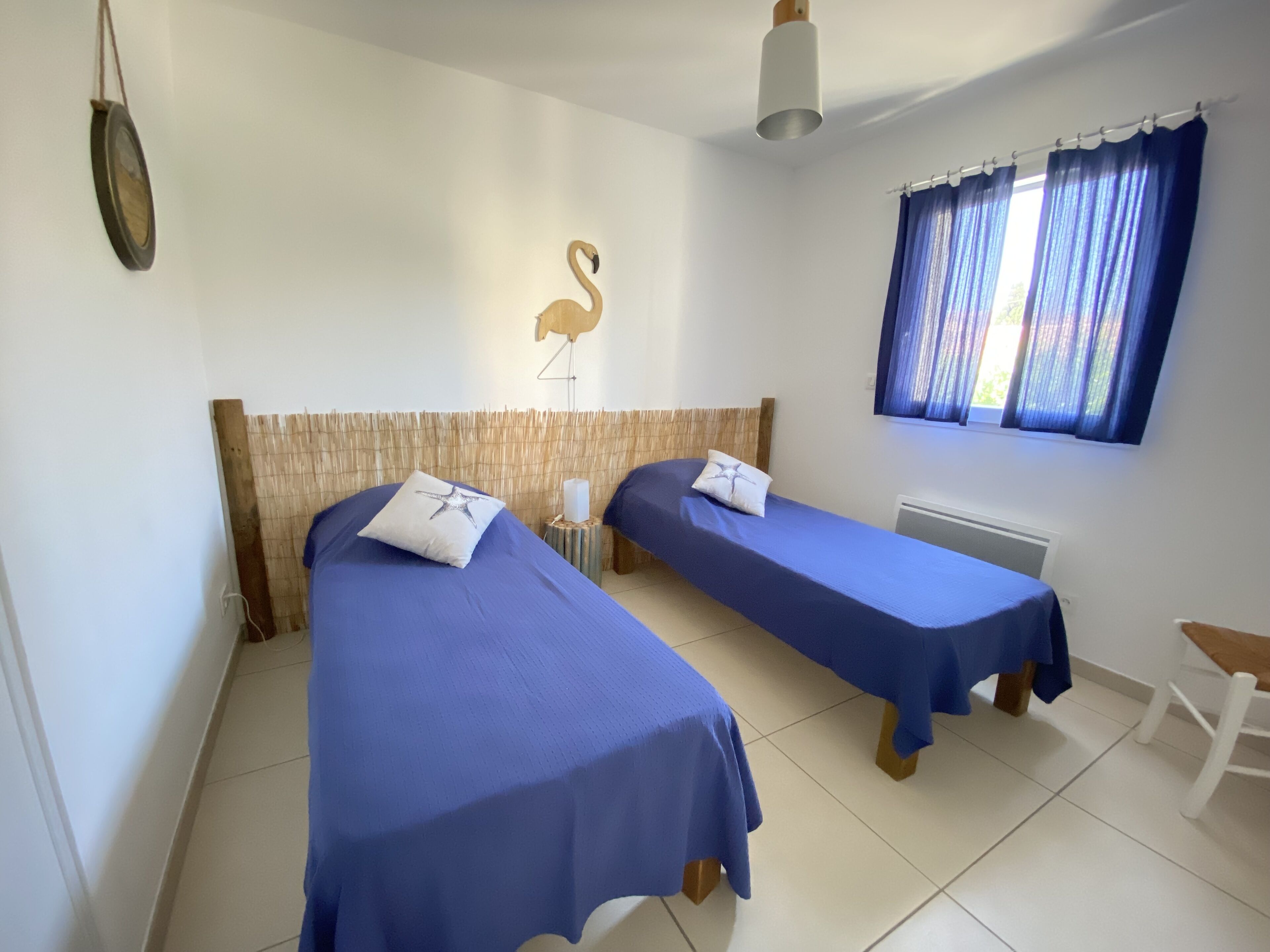 A room with two single beds, a wooden headboard, a wall-mounted light fixture, and a window with curtains.