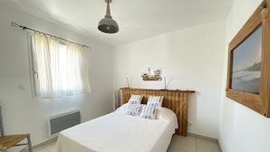 Ein Schlafzimmer mit einem hölzernen Kopfende, einem Bett mit weißer Bettwäsche, einem Fenster mit leichtem Vorhang, einem Heizkörper, einem Holzstuhl und einem an der Wand hängenden Bild.