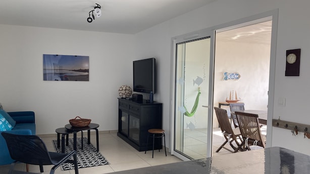 Ein modernes Wohnzimmer mit einem blauen Sofa, einem Fernseher auf einem schwarzen Schrank, einer Glastür zu einem Essbereich mit Tisch und Stühlen sowie einer wandmontierten Uhr.