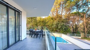 A modern outdoor area with a pool, a dining table with chairs, and a glass railing overlooking a garden.
