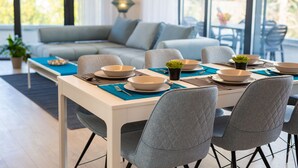 A dining area with a table set for four, surrounded by grey upholstered chairs. A sofa and a lamp are visible in the background.