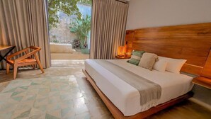 Un dormitorio con cabecera de madera, cama con ropa blanca, una silla de madera, una mesita de noche con lámpara y vista a un jardín a través de una puerta corrediza.