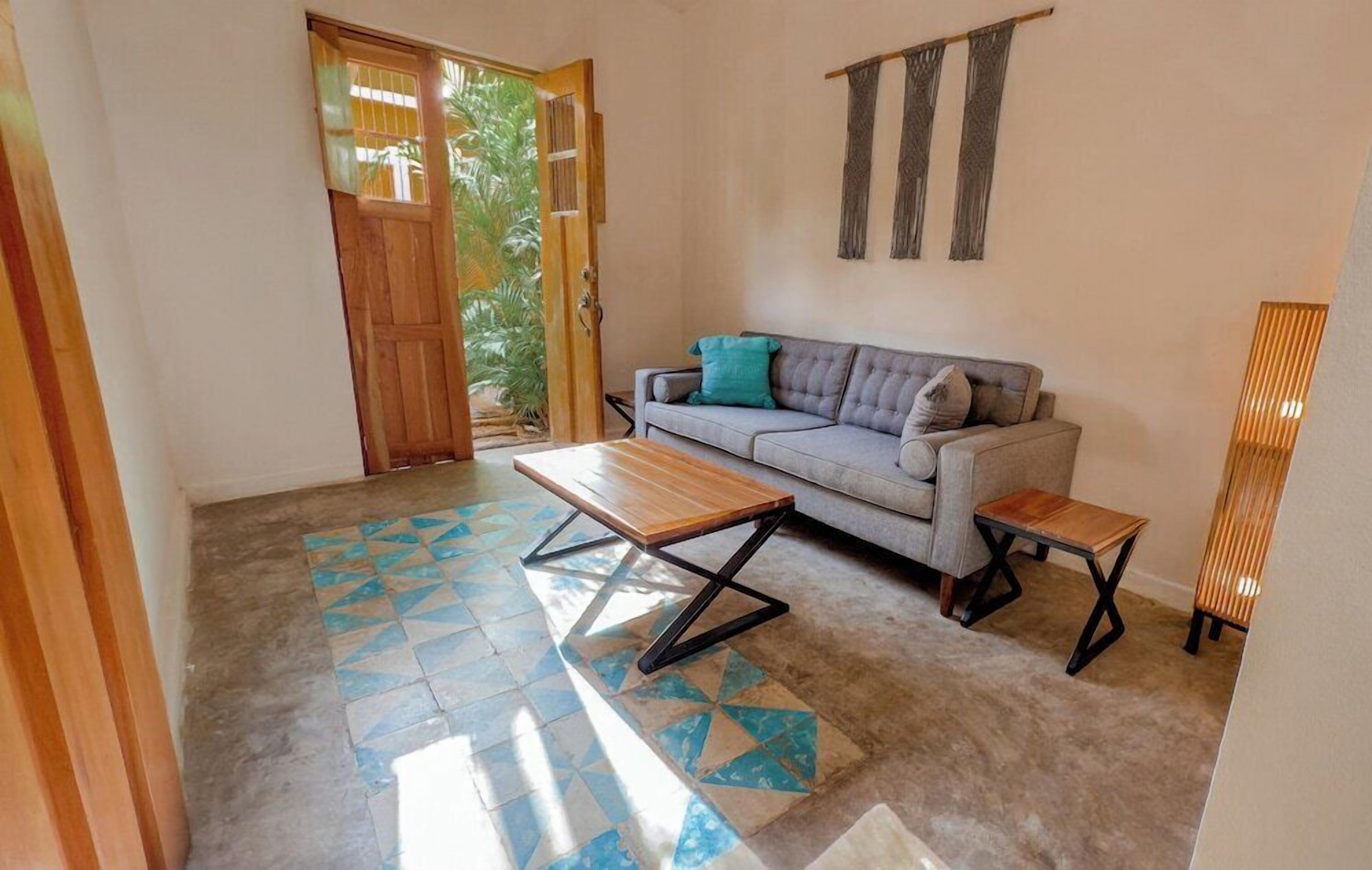 A living room with a sofa, a wooden bench, and a small table. There is a door leading to a balcony with a view of greenery.
