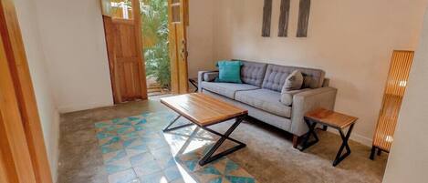 A living room with a sofa, a wooden bench, and a small table. There is a door leading to a balcony with a view of greenery.