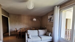 A living room with a grey sofa, a small round table, a dining area with wooden chairs, a pendant light, and a large window with curtains. - T4 apartment in the heart of Briançon, 400 m from the Serre-Chevalier slopes (Briançon)