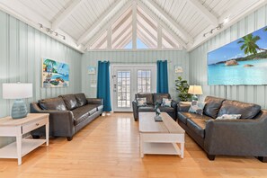 Een ruime woonkamer met een hoge plafond, twee leren banken, een salontafel en een groot schilderij van een strandgezicht.