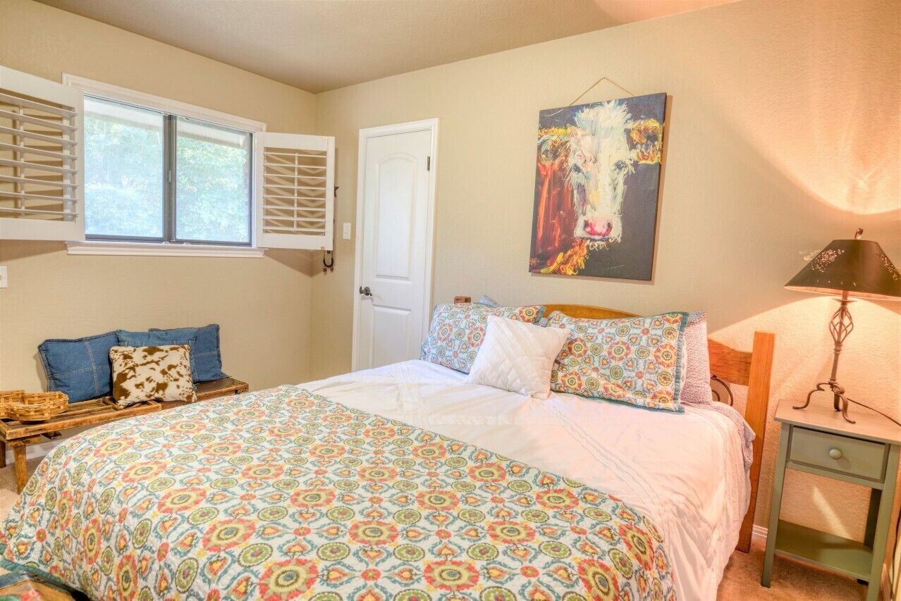 A bedroom with a bed, a window with blinds, a door, a painting, and a nightstand with a lamp.