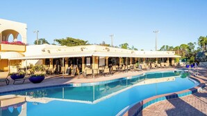 Un espace aménagé au bord de la piscine, avec des chaises longues et des parasols, une piscine, et un bâtiment doté d’une terrasse couverte.