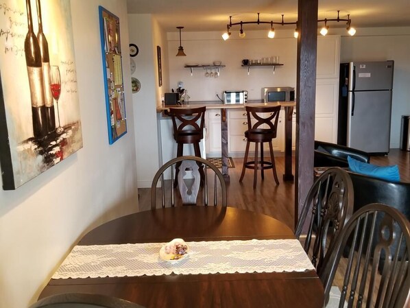 Un coin repas avec une table en bois, des chaises et un chemin de table en dentelle. Au fond, on aperçoit une cuisine équipée d’un réfrigérateur, d’un four à micro-ondes et d’une machine à café.
