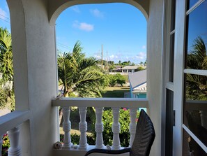 Un balcon doté de balustrades blanches, d’une chaise en osier et offrant une vue sur une piscine et la verdure environnante.