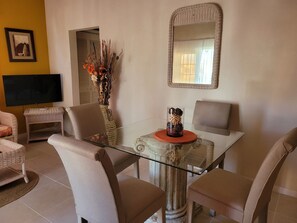Un coin repas moderne avec une table à plateau en verre, des chaises en osier, un vase contenant des fleurs séchées et un téléviseur à écran plat posé sur un meuble.