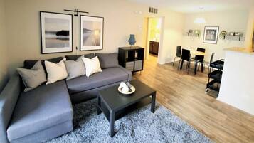 A modern living room with a grey sofa, a black coffee table, and a dining area with a kitchen in the background.