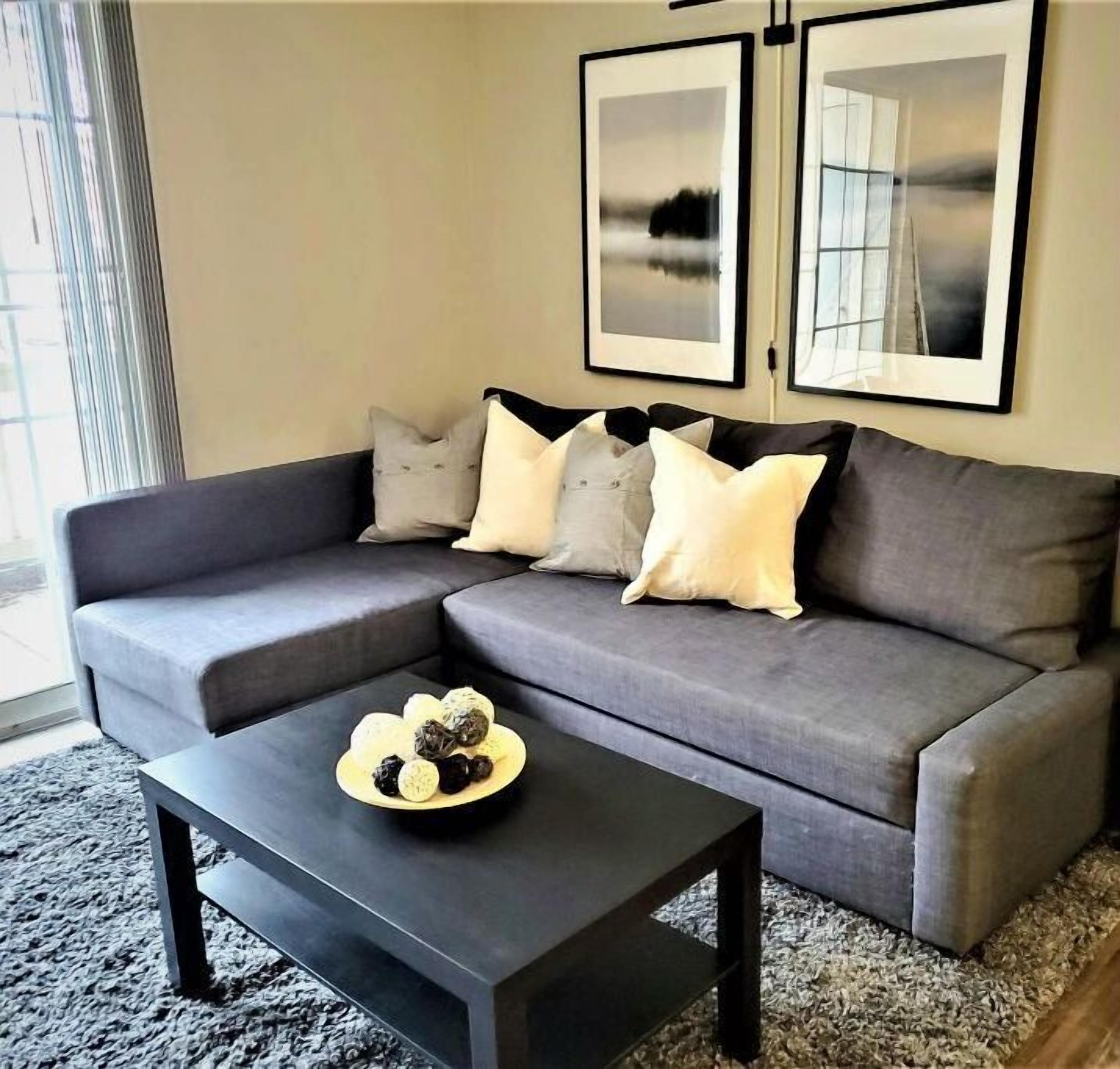 A modern living room with a grey sectional sofa, a black coffee table, and a rug. There are three framed pictures on the wall.