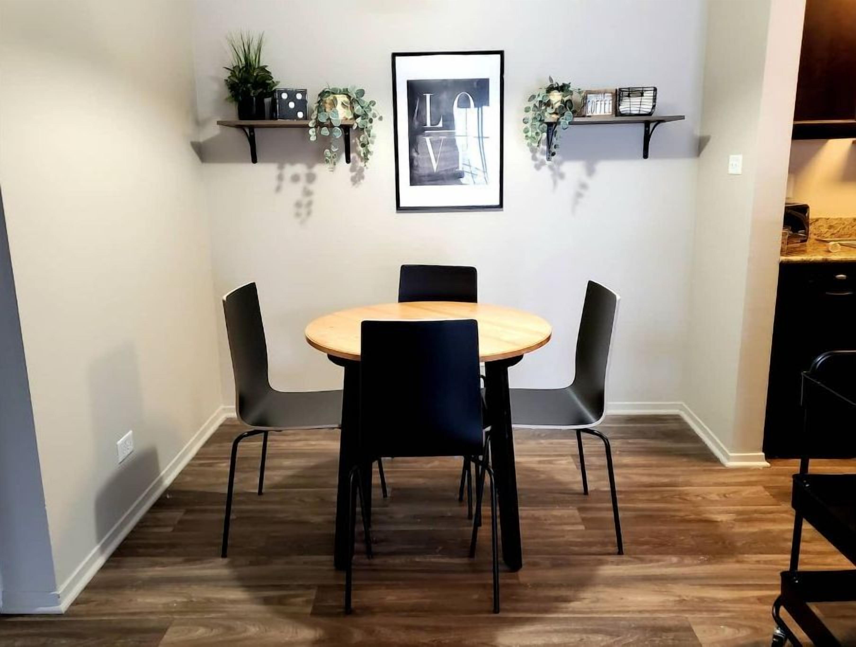 A dining area with a round wooden table and four chairs, flanked by two shelves with plants and a framed picture.