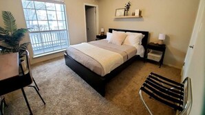 A bedroom with a bed, bedside tables, a desk, a chair, a ceiling fan, a window, and a door.