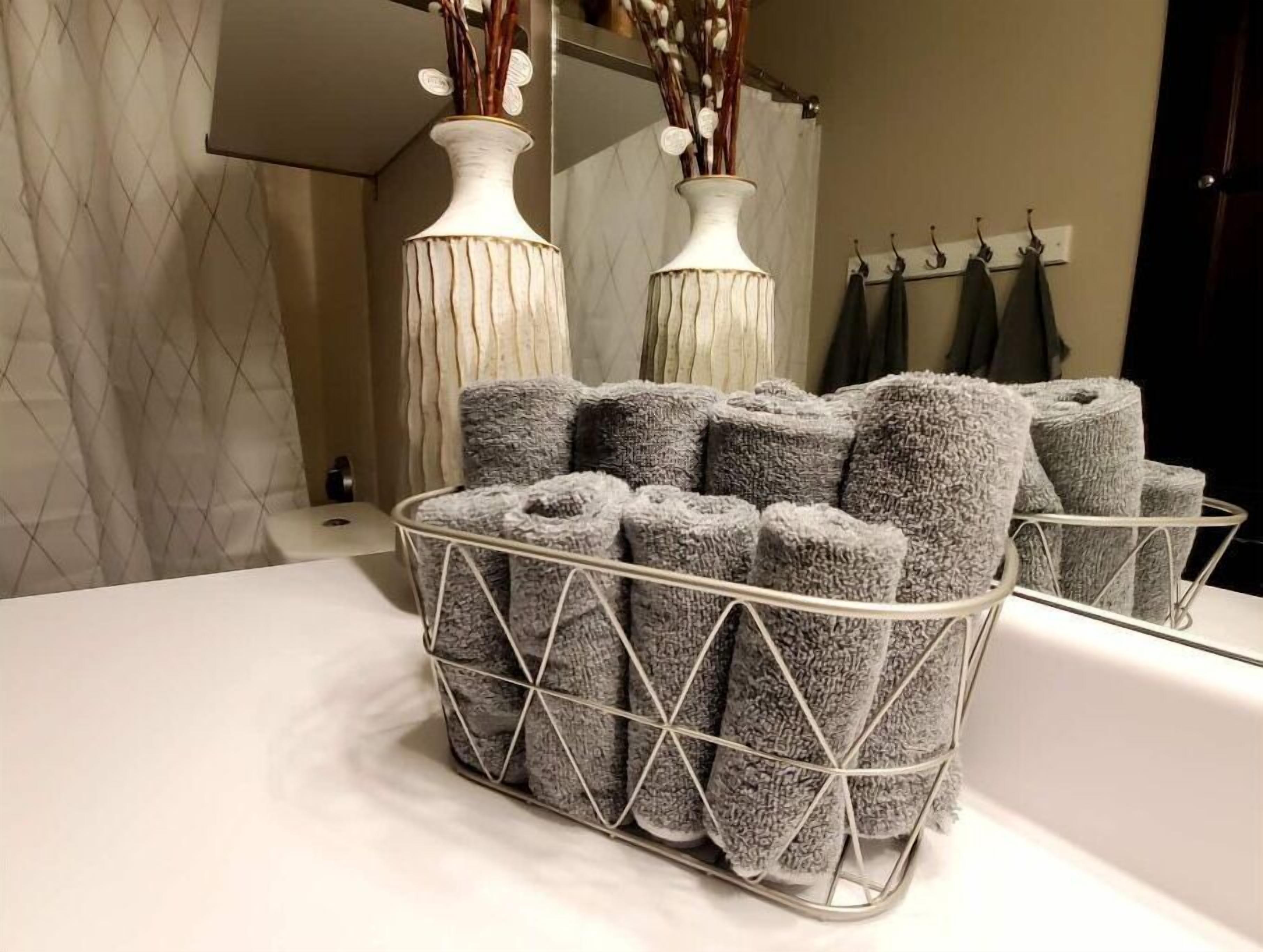 A bathroom with a white sink, two white vases with dried branches, and a metal basket holding rolled towels.
