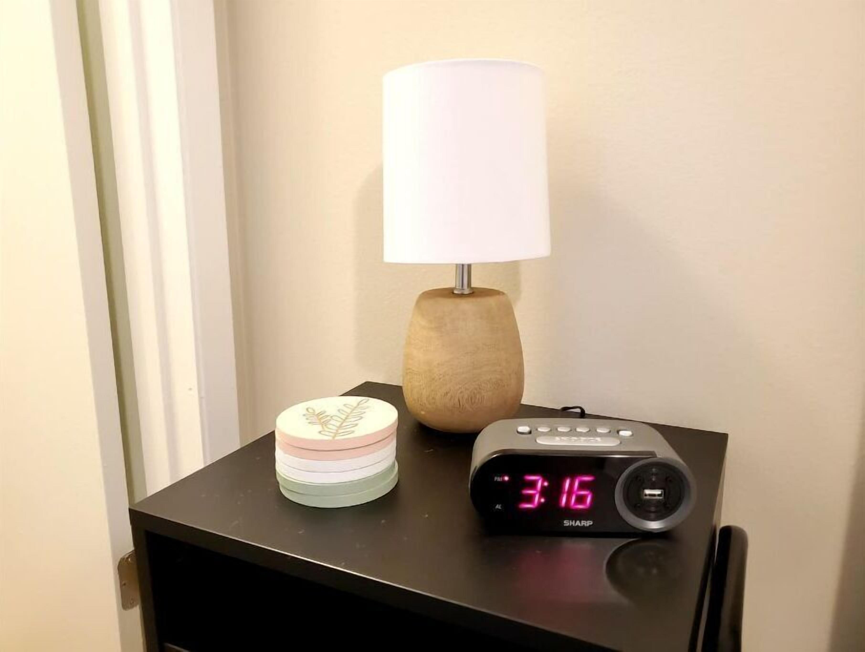 Een nachtkastje met een digitale klok waarop 15:16 staat, een lamp en een decoratieve pot.