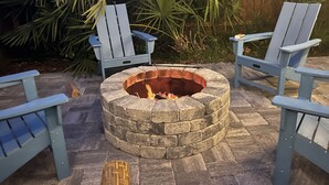 A stone fire pit with two Adirondack chairs and a wooden table.