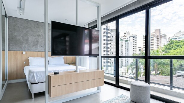 A modern bedroom with a bed, a TV, a wooden dresser, and a large window offering a city view. - ZEE Studios by Viva - Studios a 200m da beira-mar norte (Florianópolis)
