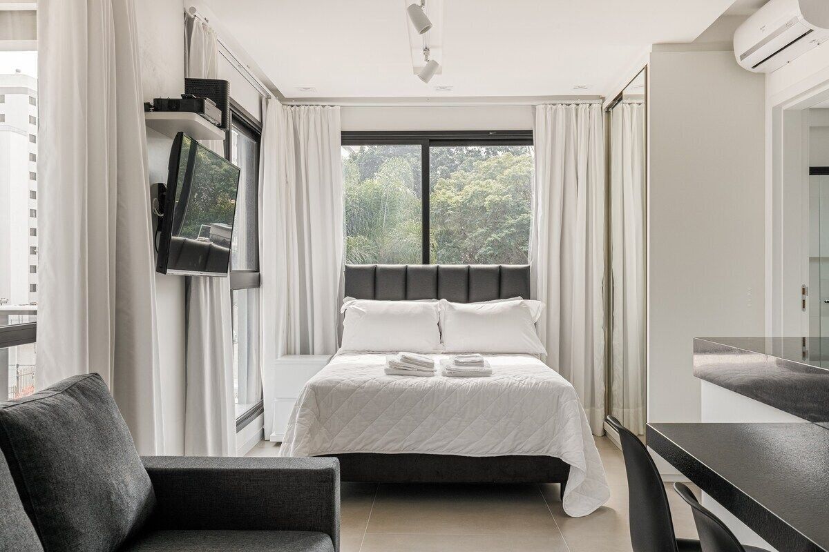 A modern hotel room with a bed, a TV mounted on the wall, a desk, and a window with curtains.