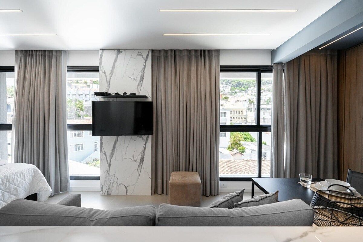 Una habitación de hotel moderna con un ventanal grande, un televisor de pantalla plana montado en una pared de mármol, un sofá gris y un comedor con una mesa preparada para dos personas.