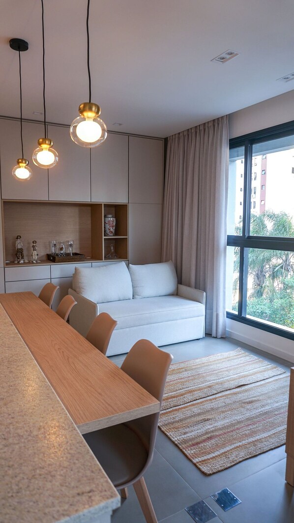 A modern living room with a sofa, dining table, and chairs. There are pendant lights, a large window with curtains, and a view of buildings outside. - ZEE Studios by Viva - Studios a 200m da beira-mar norte (Florianópolis)