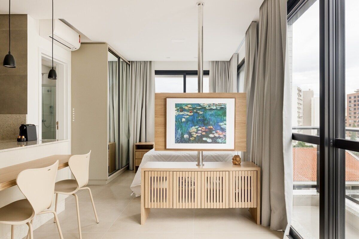A modern hotel room with a wooden console table, a framed painting, a bed, a desk with chairs, and large windows offering a city view.