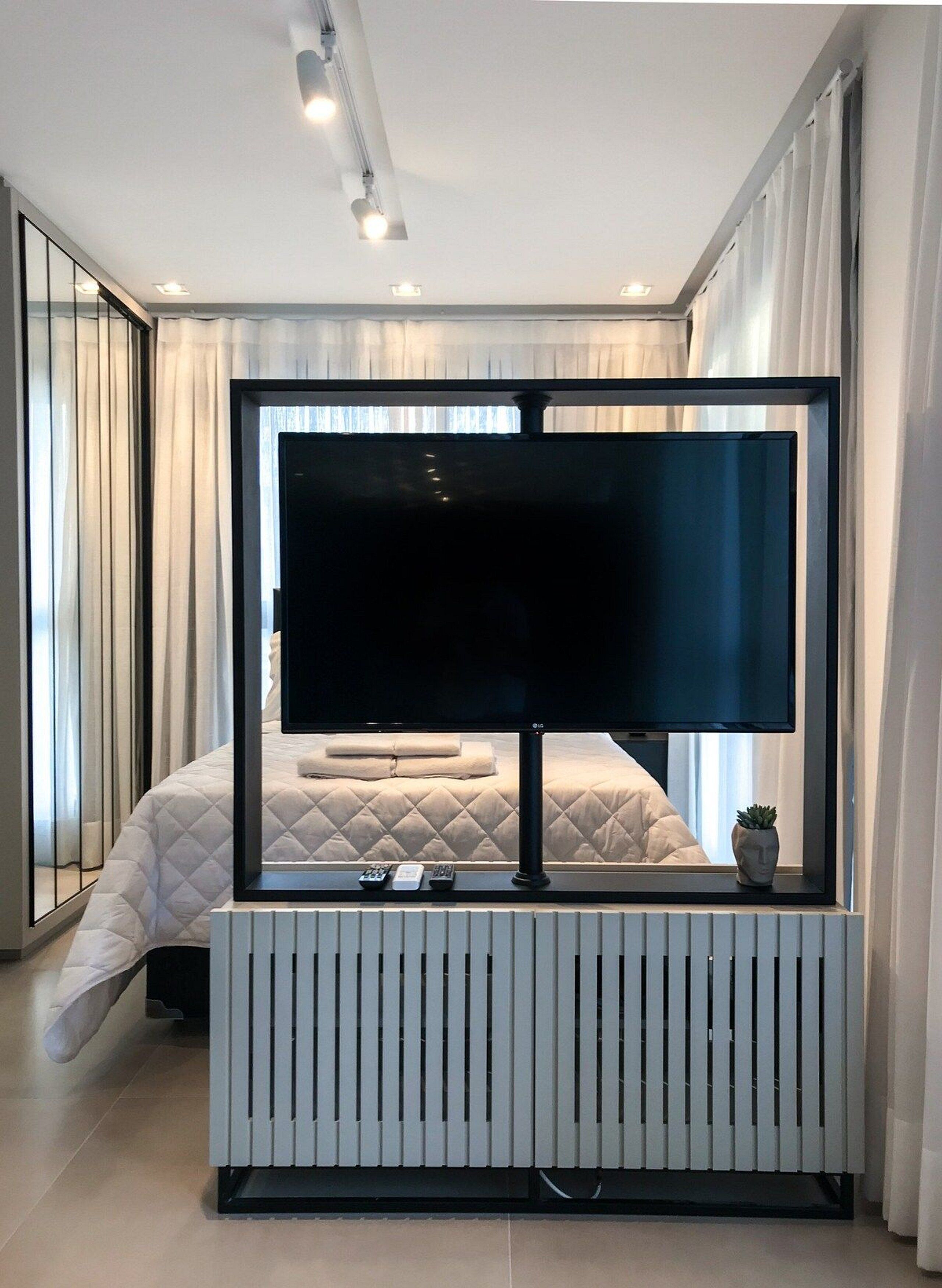Ett modernt hotellrum med en stor platt-TV som är monterad på ett vitt element, en säng med ett vadderat täcke och en krukväxt på fönsterbrädan.