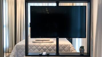 Ett modernt hotellrum med en stor platt-TV som är monterad på ett vitt element, en säng med ett vadderat täcke och en krukväxt på fönsterbrädan.