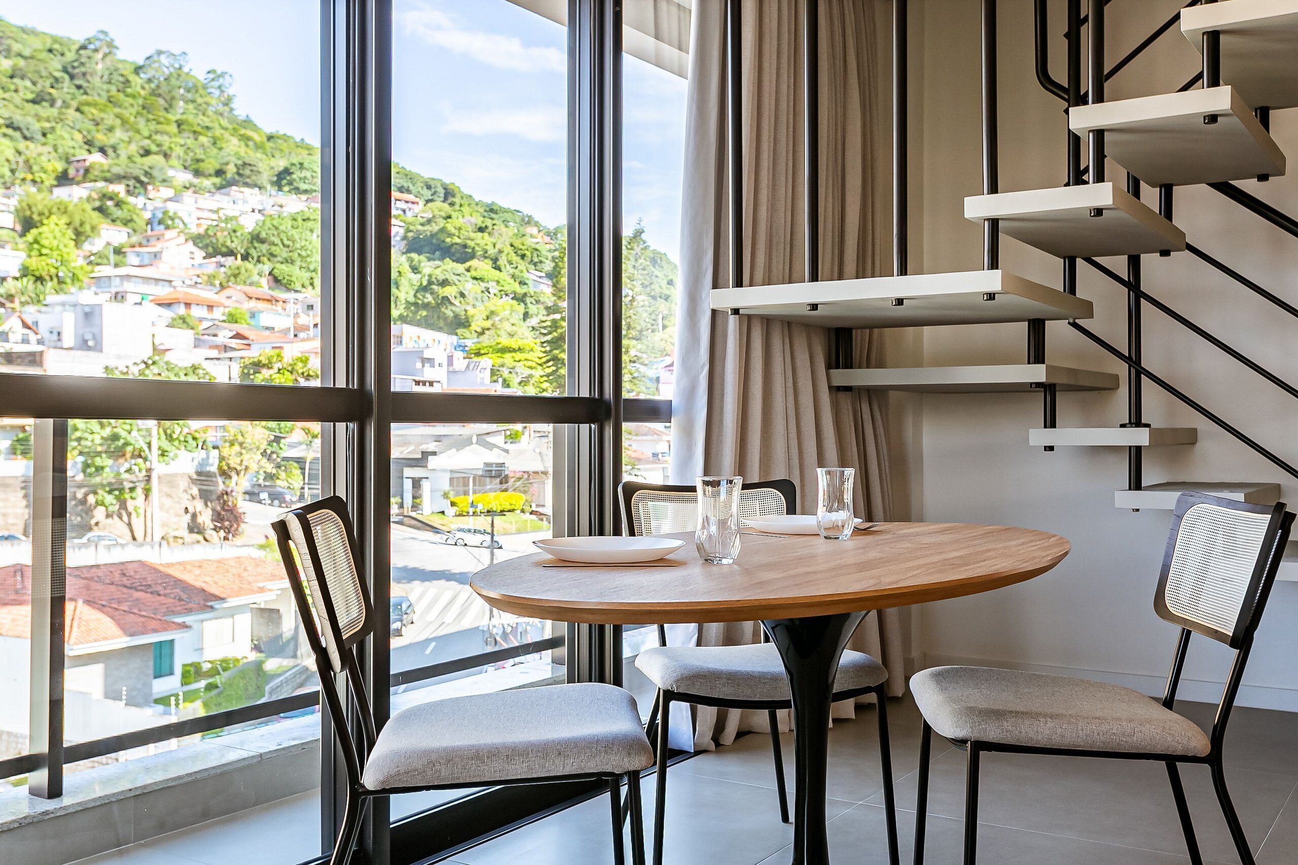 A modern dining area with a round table, two chairs, and a view of a hillside town through large windows.