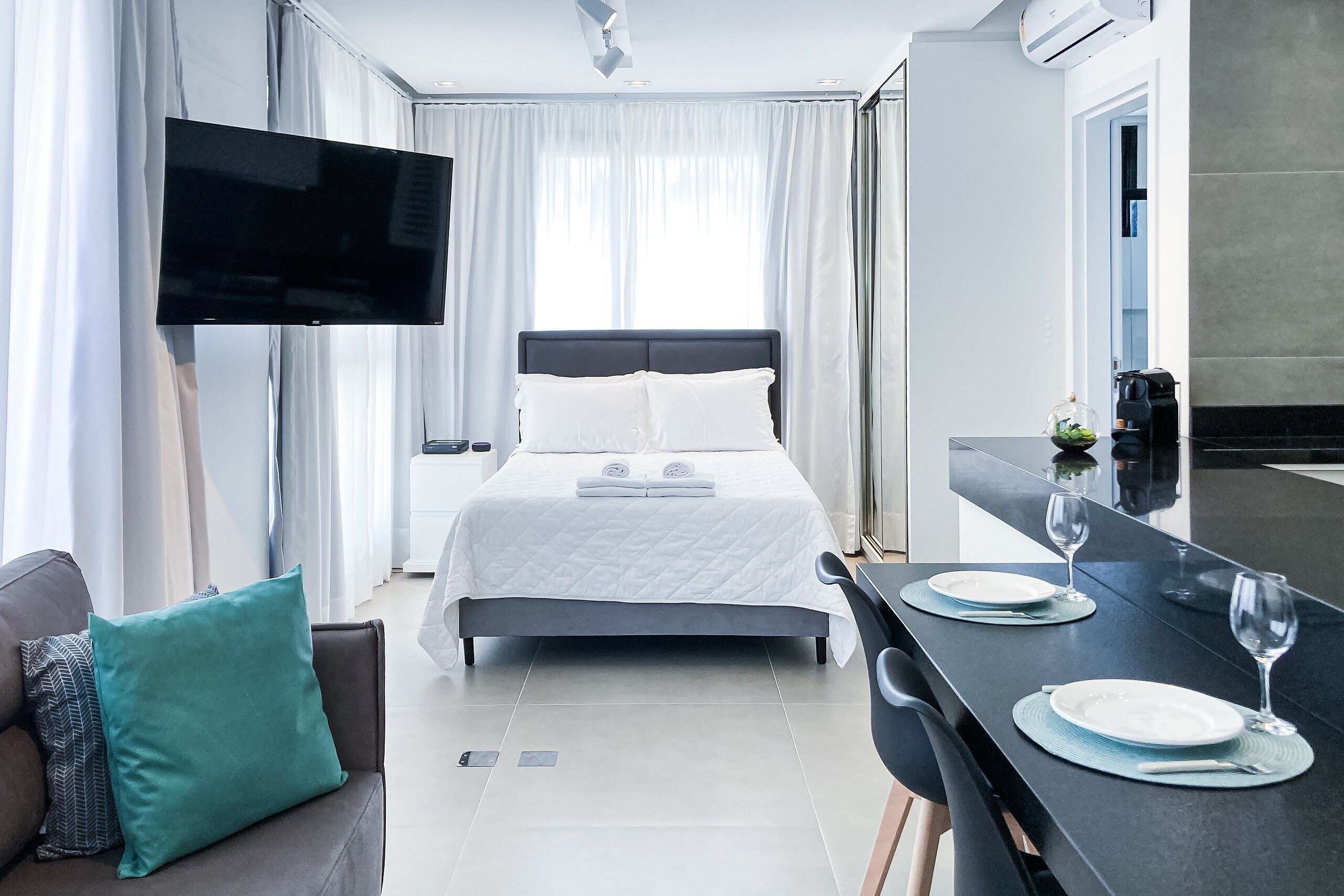 A modern hotel room with a bed, a TV mounted on the wall, a dining table with plates and glasses, and a sofa with cushions.