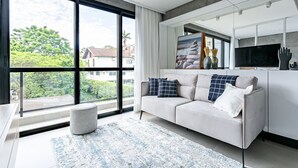 A modern living room with a sofa, a rug, and a large window offering a view of greenery and buildings. - ZEE Studios by Viva - Studios a 200m da beira-mar norte (Florianópolis)