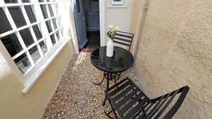 Een klein buitgedeelte met een ronde tafel en twee stoelen, een witte vaas met bloemen, en een deur die leidt naar een blauwe kamer.