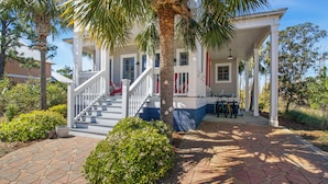A covered porch with a staircase leading to a second floor, surrounded by palm trees and a brick pathway. - THE GOOD STUFF IS A LUXUROUS 3BD HOME W/COMMUNITY POOL, GATED COMMUNITY! (Port Saint Joe)