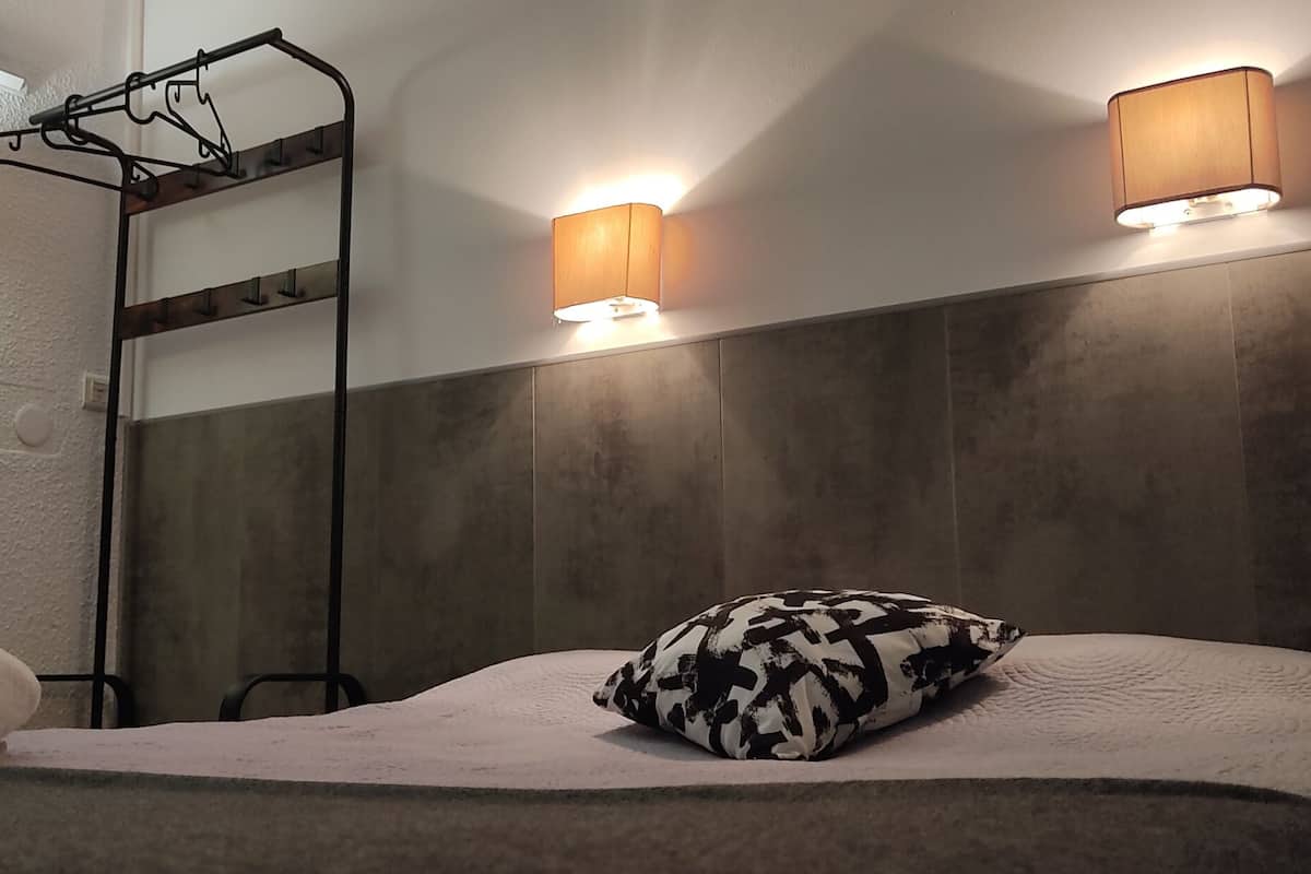 A neatly made bed with a patterned pillow and a rolled towel, a metal rack with hooks, and two wall-mounted lamps.