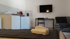 Quarto compacto com cama, escrivaninha, cadeira, televisão e cozinha compacta.