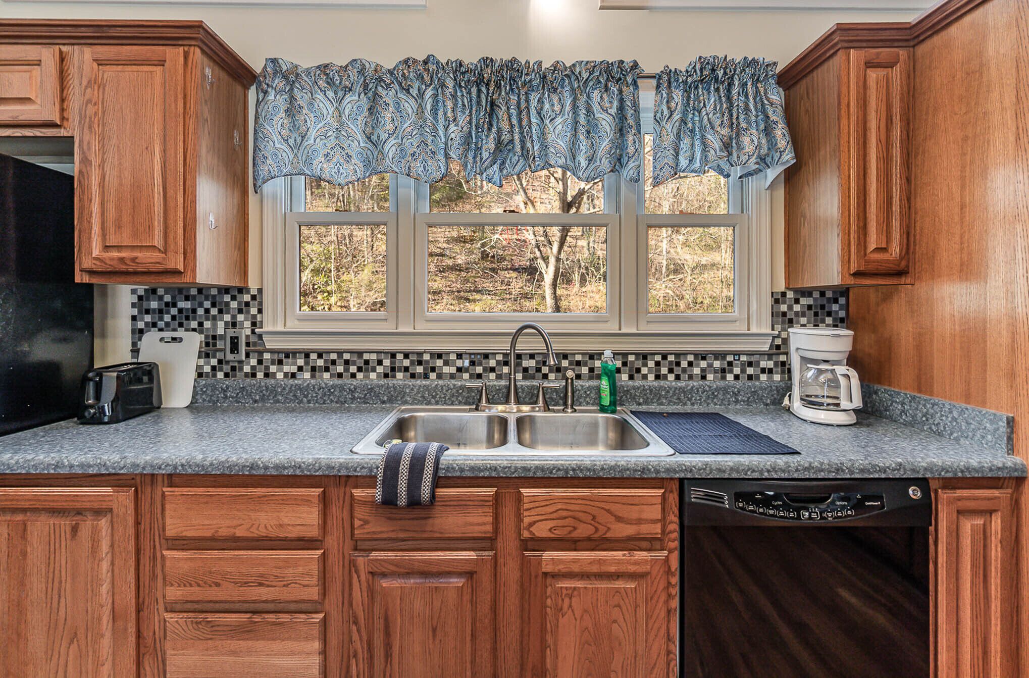 A kitchen with wooden cabinets, a double sink, a coffee maker, and a window with a view of trees.
