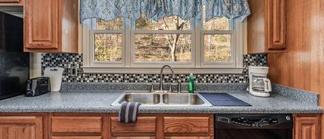 A kitchen with wooden cabinets, a double sink, a coffee maker, and a window with a view of trees.