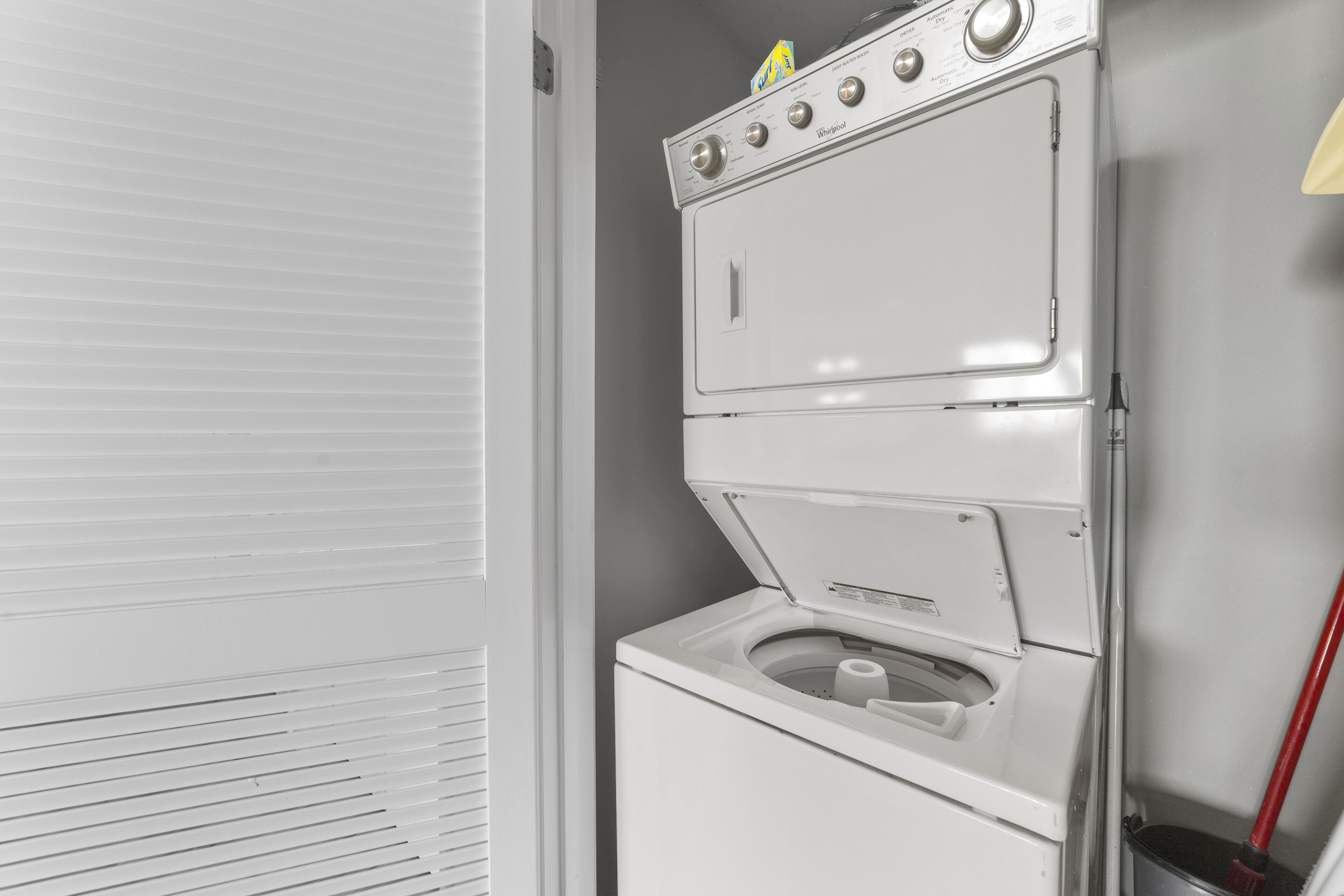 A stacked washer and dryer unit with a control panel and a red broom leaning against the wall.