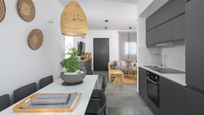A modern kitchen with dark cabinets, a white countertop, and a built-in oven. There is a dining area with a white table and chairs, a large plant, and a hanging light fixture. - Seasand Villa (Rhodes)
