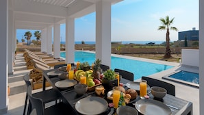 A dining area with a pool view, featuring a table set with breakfast items and a tropical fruit bowl. - Seasand Villa (Rhodes)