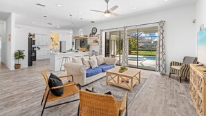A spacious living area with a sofa, coffee table, and wicker chairs. A kitchen is visible in the background, and there is a large sliding glass door leading to a pool area. -  Large pool/spa home minutes from beach with arcade games and canal for fishing. (Bradenton)