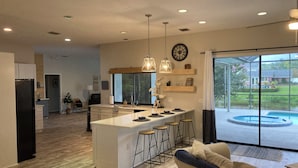 A modern kitchen with a central island, bar stools, pendant lighting, and a large window overlooking a pool. -  Large pool/spa home minutes from beach with arcade games and canal for fishing. (Bradenton)