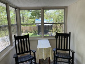 Ein Wintergarten mit zwei Holzstühlen und einem Tisch, der auf einen Garten mit Holzzaun und Gartenhaus blickt.