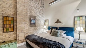 A bedroom with a brick wall, a bed with a blue headboard, a small bedside table, and a green trunk. - Cozy 1BD w/ View in Walker's Point, Free Parking (Milwaukee)