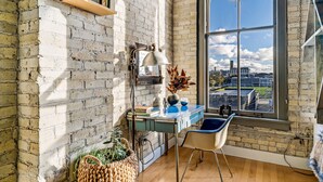 A room with a brick wall, a large window with a view of the city, a desk with a lamp, and a chair. - Cozy 1BD w/ View in Walker's Point, Free Parking (Milwaukee)