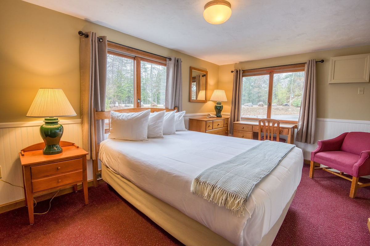 A hotel room with a large bed, two bedside tables, a desk, a chair, and a window with curtains.